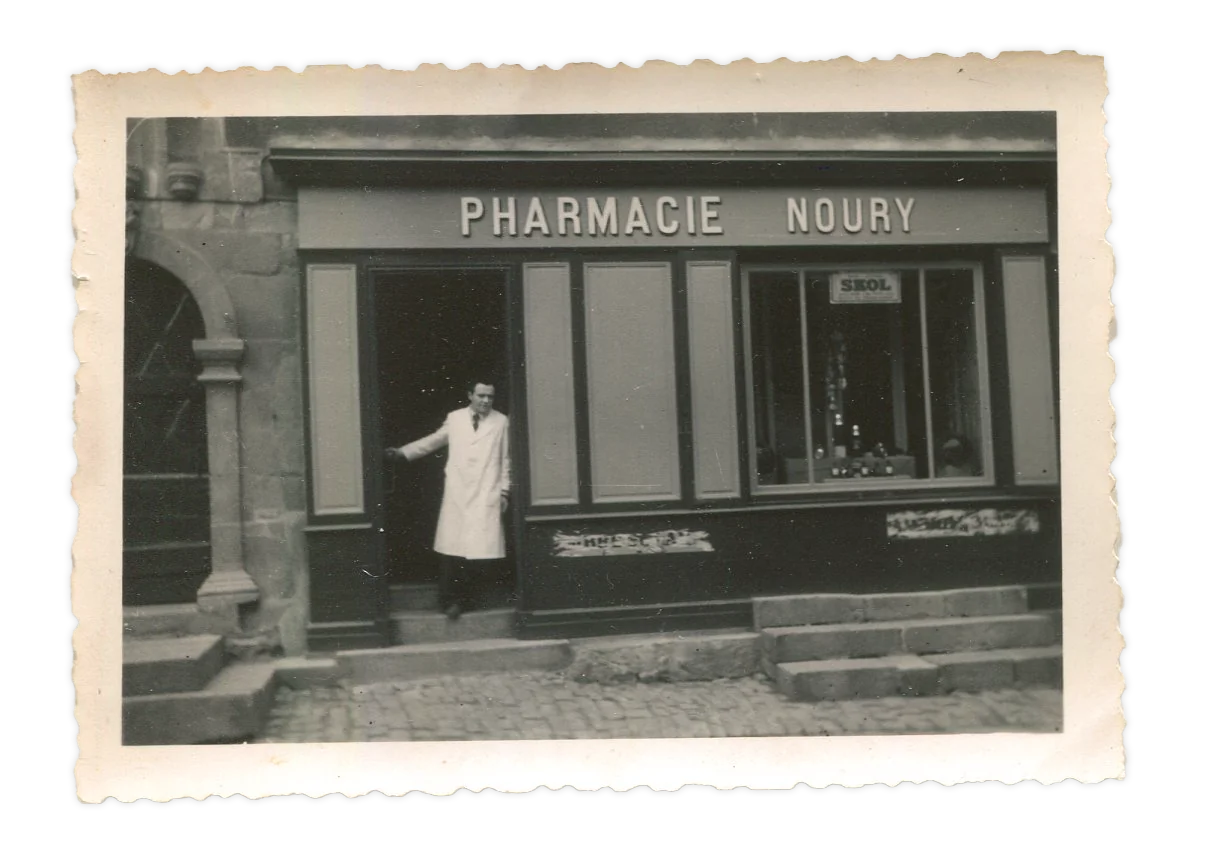 Photo noir et blanc de la devanture Pharmacie Noury, avec le pharmacien debout sur le seuil